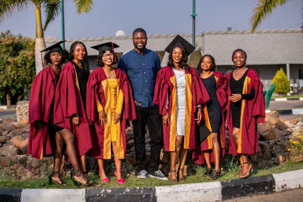 6 graduating girls wearing their caps and gowns and smiling with Lord Mbambu in the middle.