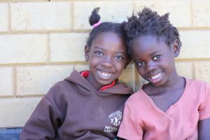 Two Zambian girls sitting in front of a brick wall and smiling together.