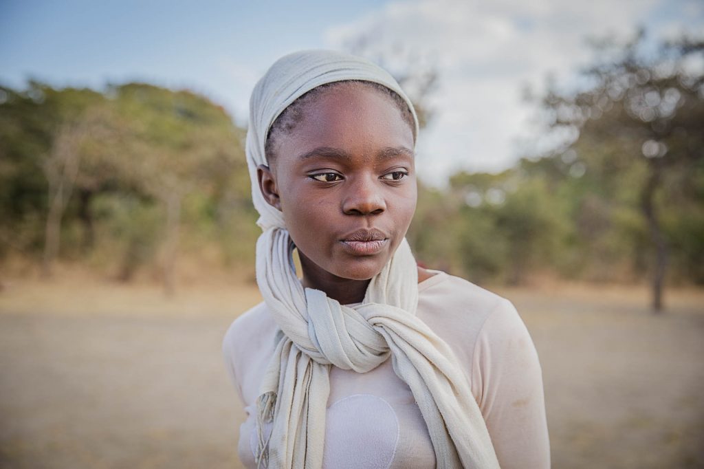 A Zambian girl wearing a head scarf standing in front of Acacia trees.