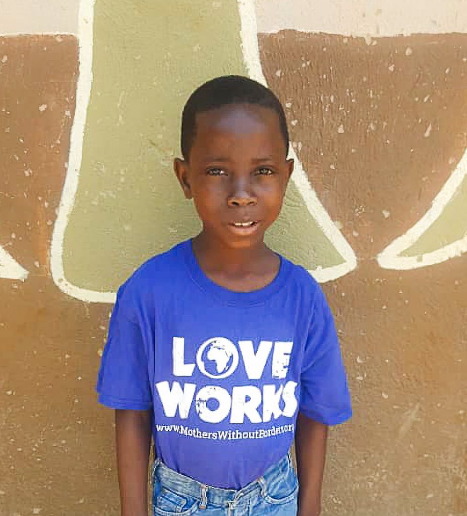 Little Zambian boy standing against a wall and wearing a "love works" t-shirt.