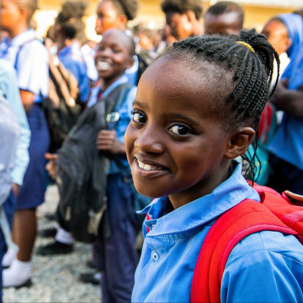Zambian school girl smiling and wearing a red backpack with other school kids in the background.